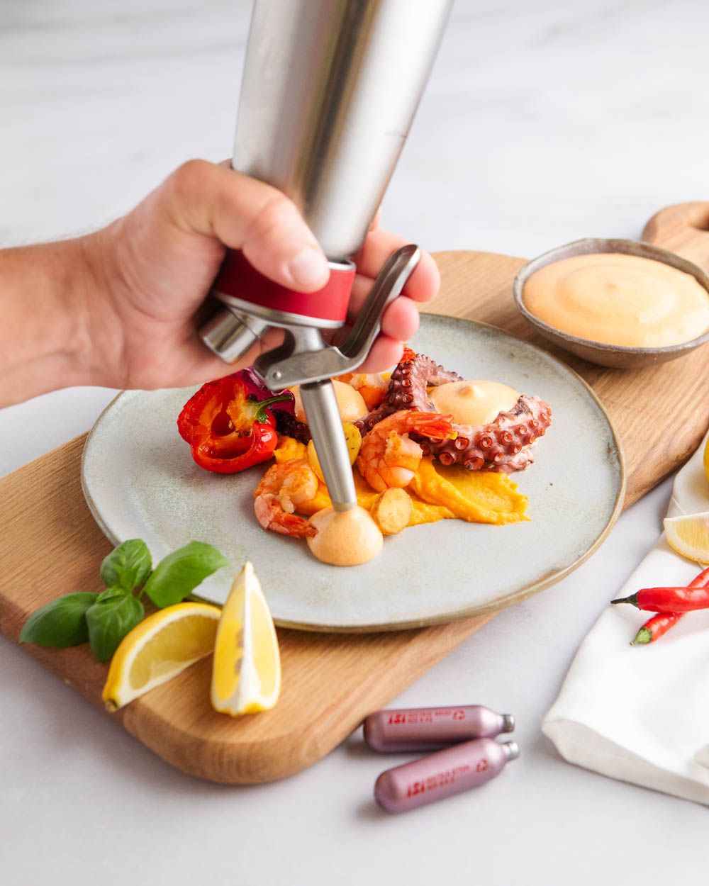 Jemand nutzt einen iSi Gourmet Whip um Chilli Mayonnaise auf einen Teller mit Meeresfrüchten zu sprühen. Daneben liegen Zitronen und zwei iSi Charge Kapseln. 