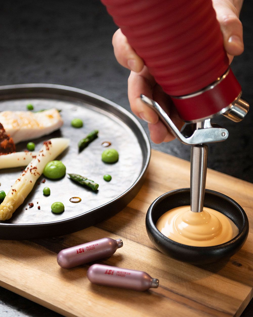 Auf dem Bild sieht man, wie mit einem iSi Gourmet Whip eine helle Sauce in eine schwarze Schale gespritzt wird, daneben liegt ein Teller mit Gemüse.