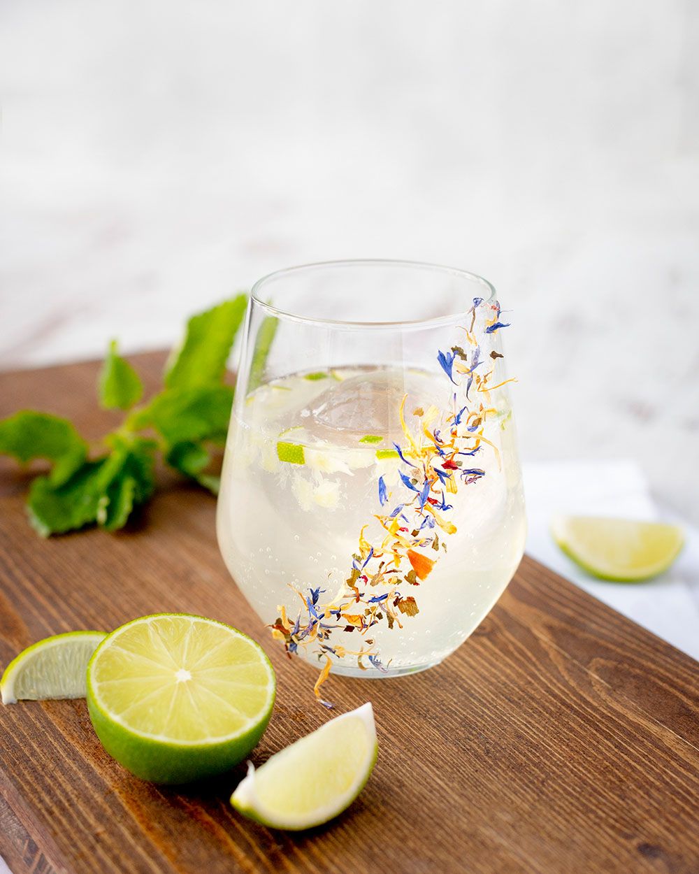 Ein Glas mit sprudeligem Holundersaft, davor Limetten, dahinter Kräuter. 