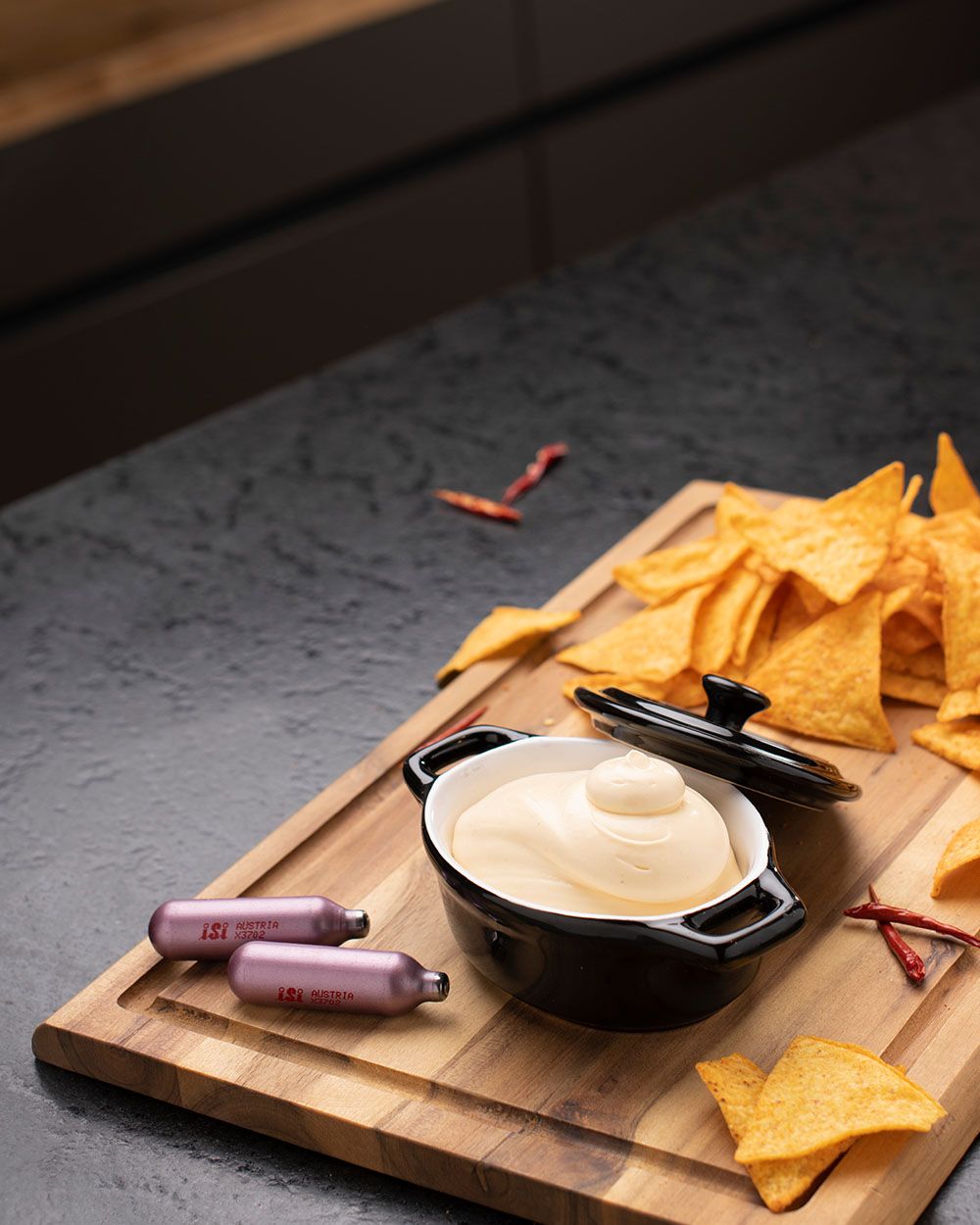 Auf dem Bild ist eine kleine schwarze Schale mit cremigem Käse-Dip zu sehen, daneben liegen Nachos auf einem Holzbrett. Zwei iSi Sahnekapseln (N₂O) sind dekorativ daneben platziert.