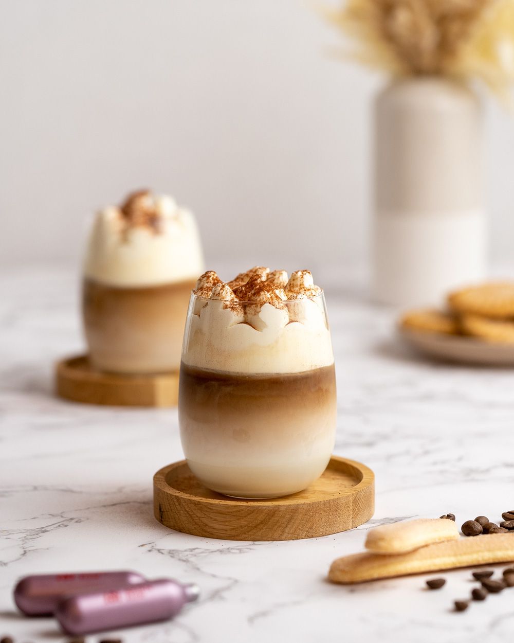 Zwei Kaffeegetränke mit Tiramisu-Topping, davor Biskotten und iSi Charge Kapseln. 