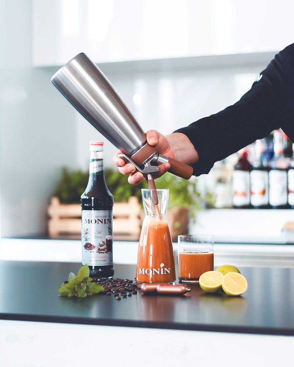 Jemand sprüht mit einem iSi Nitro Schaum auf einen Hibiscus Kombucha, daneben Monin Sirup, Zitronen, iSi Charge Kapseln und Kaffeebohnen. 