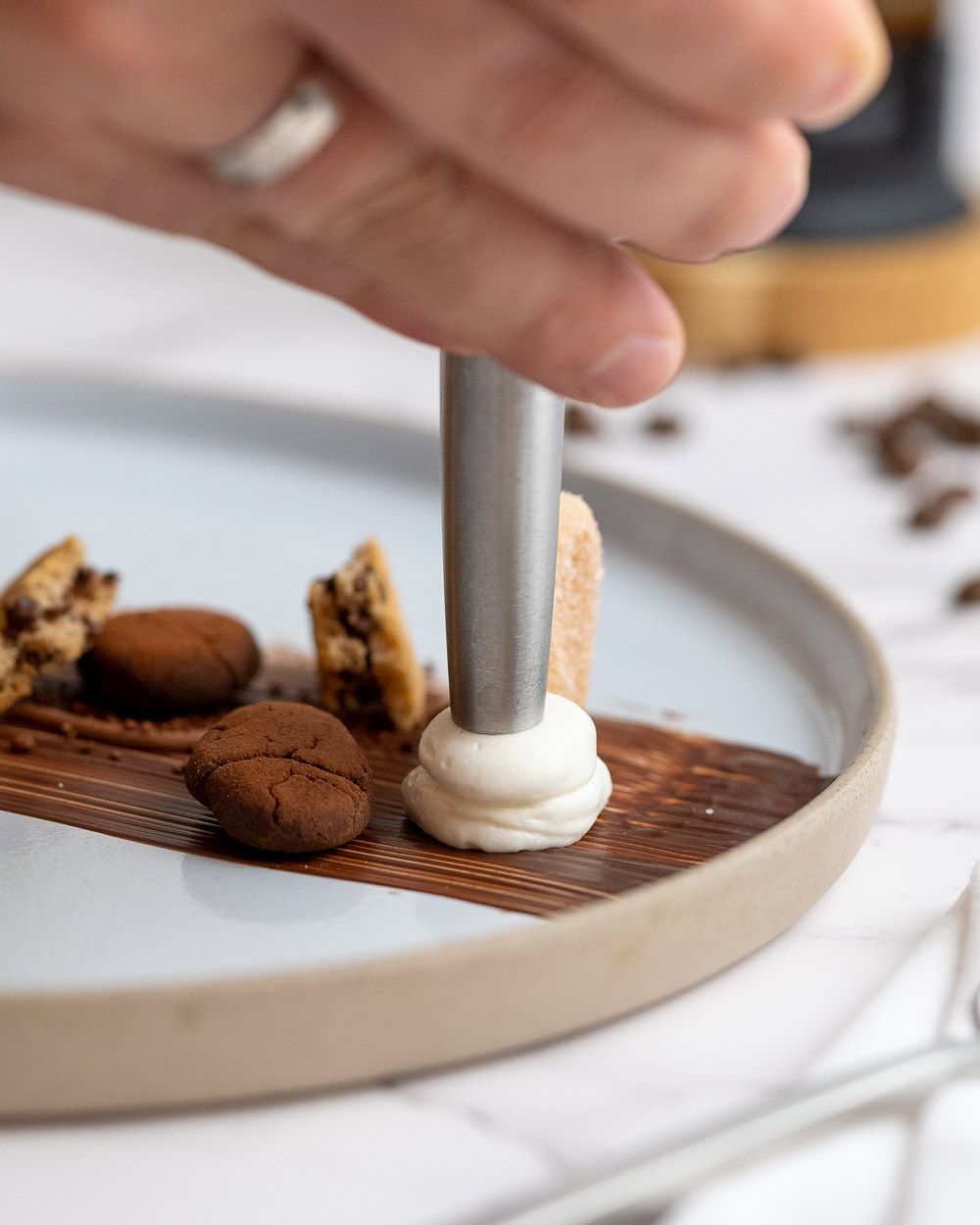 Jemand sprüht mit einem iSi Whip Schaum auf einen Teller, daneben sind Kaffeebohnen mit Kakao überzogen. 