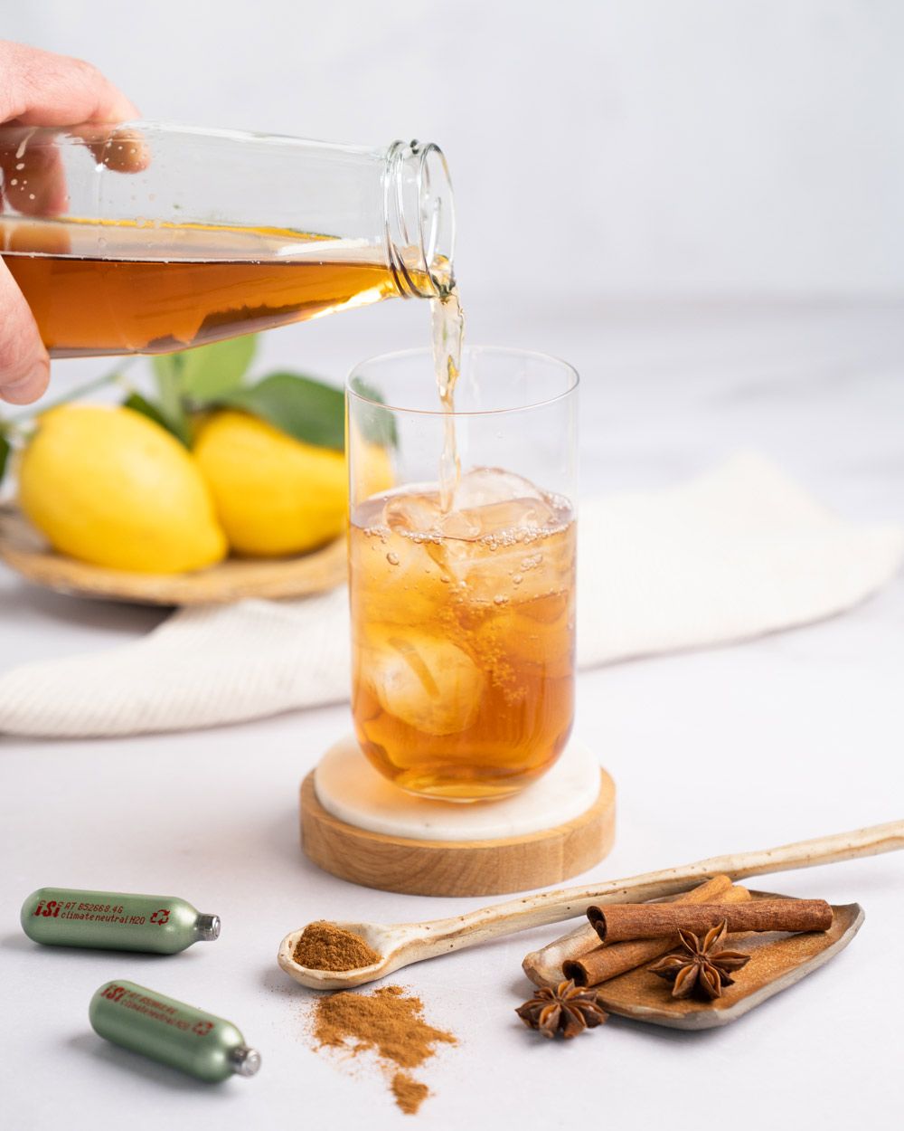 Jemand gießt Eistee in ein Glas mit Eis, daneben Zimtstangen, iSi Charge Kapseln und Zitronen. 