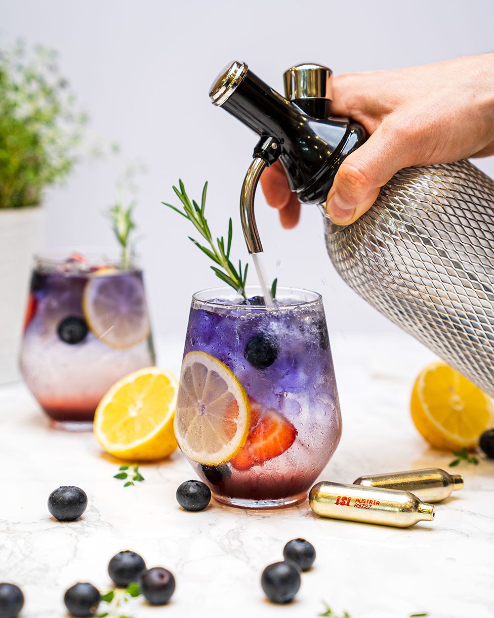 Eine Hand füllt mit einem iSi Sodamaker einen lila Cocktail mit Eiswürfeln, Zitrone, Beeren und Rosmarin, daneben liegen Zitronenhälften und goldene iSi Kapseln.