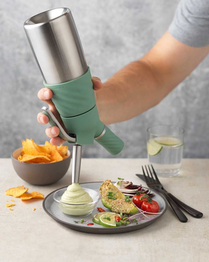 Jemand sprüht mit einem iSi Creative Whip Guacomole Dip in eine Schüssel. Daneben liegt Gemüse, im Hintergrund sind ein Glas Wasser und eine Schüssel mit Nachos zu sehen. 