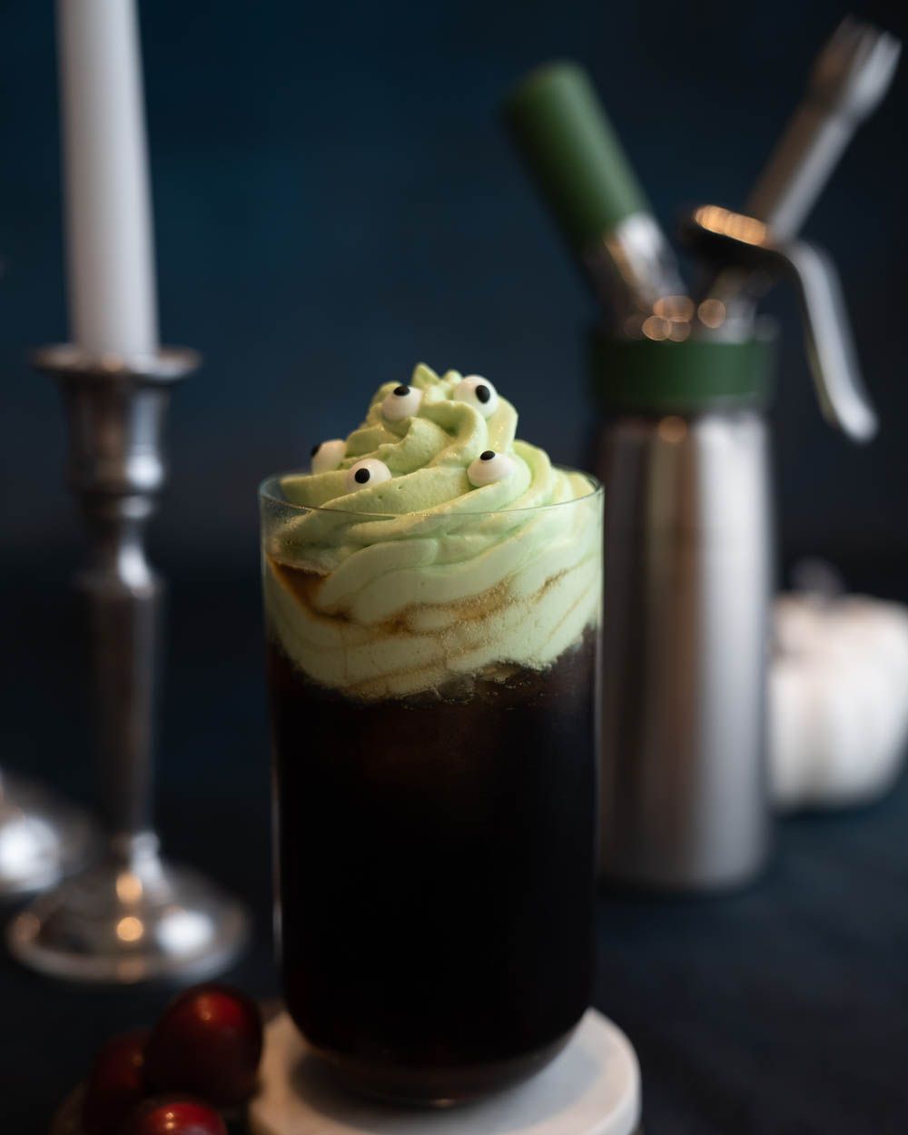 Ein Glas mit einem Kaffeegetränk, auf dem grünes Sour Apple Topping ist, im Hintergrund ein iSi Creative Whip. 