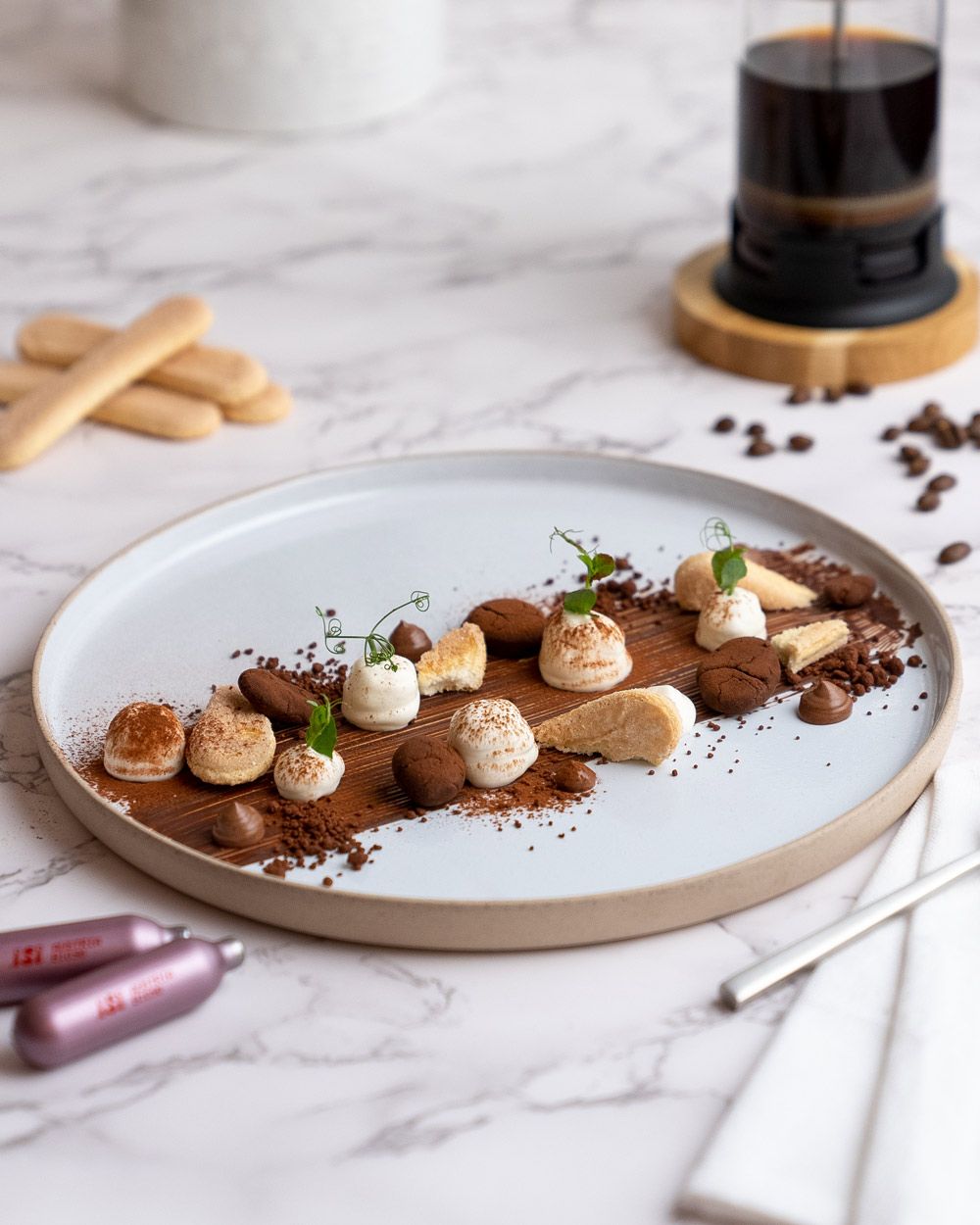 Ein Teller mit Tiramisu, daneben iSi Charge Kapseln, im Hintergrund Kaffee und Biskotten. 