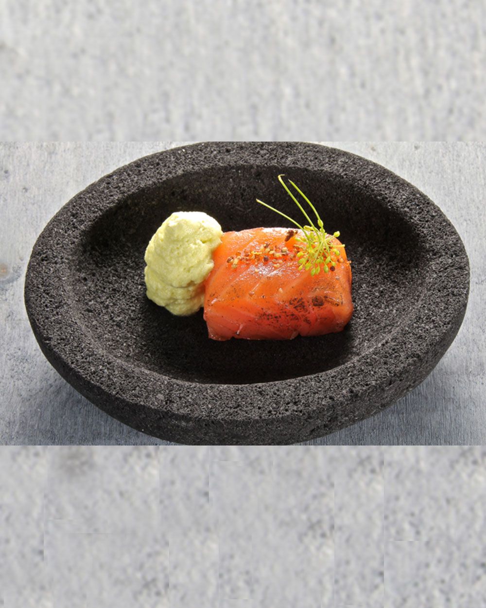 Ein Stück gebeizter Lachs ist mit Dill und einer cremigen Beilage in einem schwarzen Steinteller angerichtet.