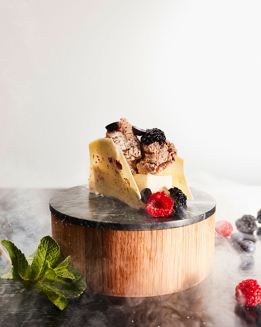 Elegantes Dessert aus weißer Schokolade mit Beeren und Schokoladencreme, dekorativ auf einem runden Holzpodest mit Minzblättern und frischen Beeren angerichtet.