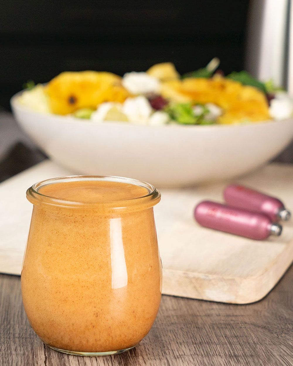 Auf dem Bild steht ein Glas mit orangefarbenem Dressing im Vordergrund, im Hintergrund ist eine Schüssel mit Salat und essbaren Blüten zu sehen.