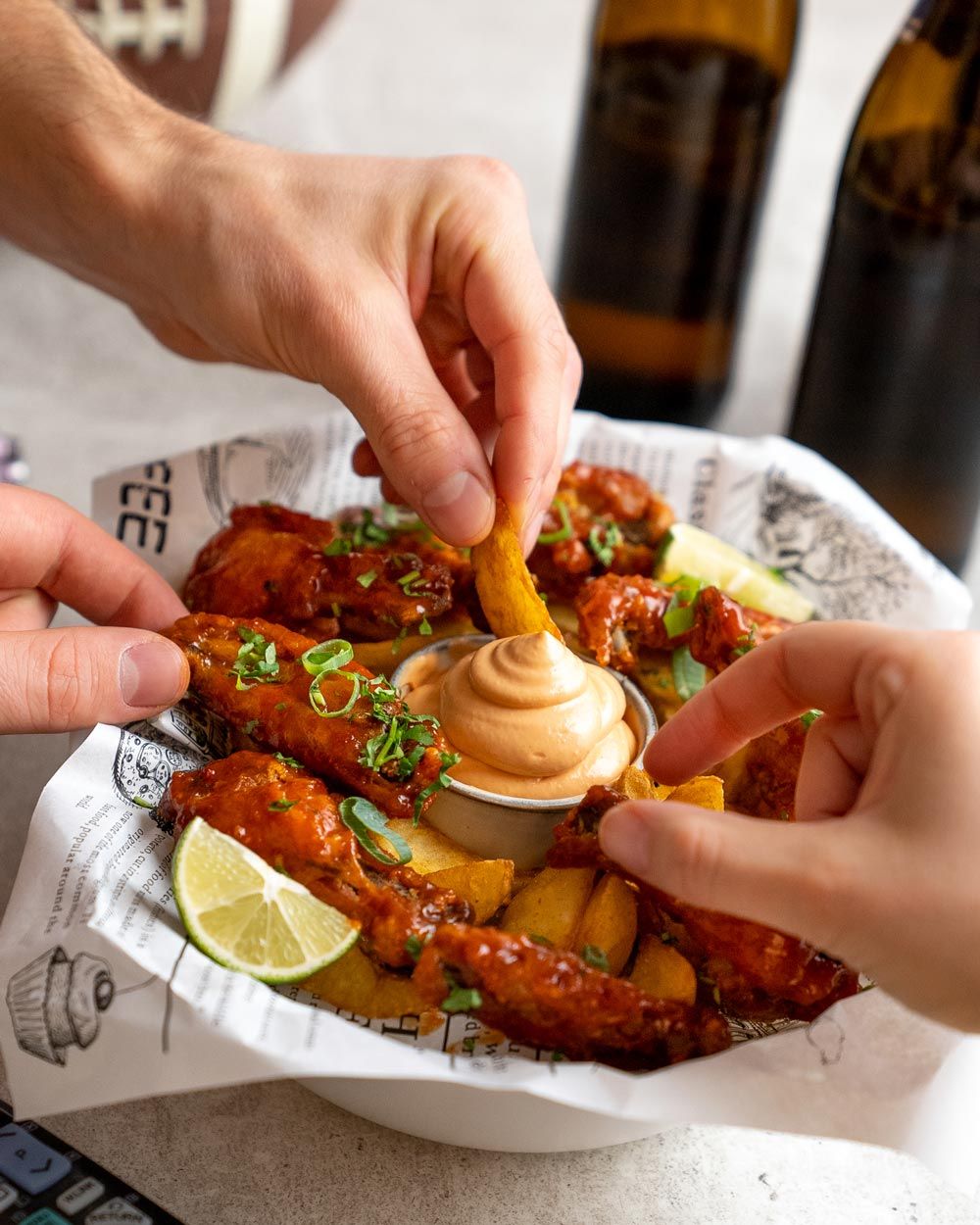 Auf dem Bild tunken zwei Hände Kartoffelspalten in einen Dip, umgeben von Chicken Wings und Zitronenspalten.