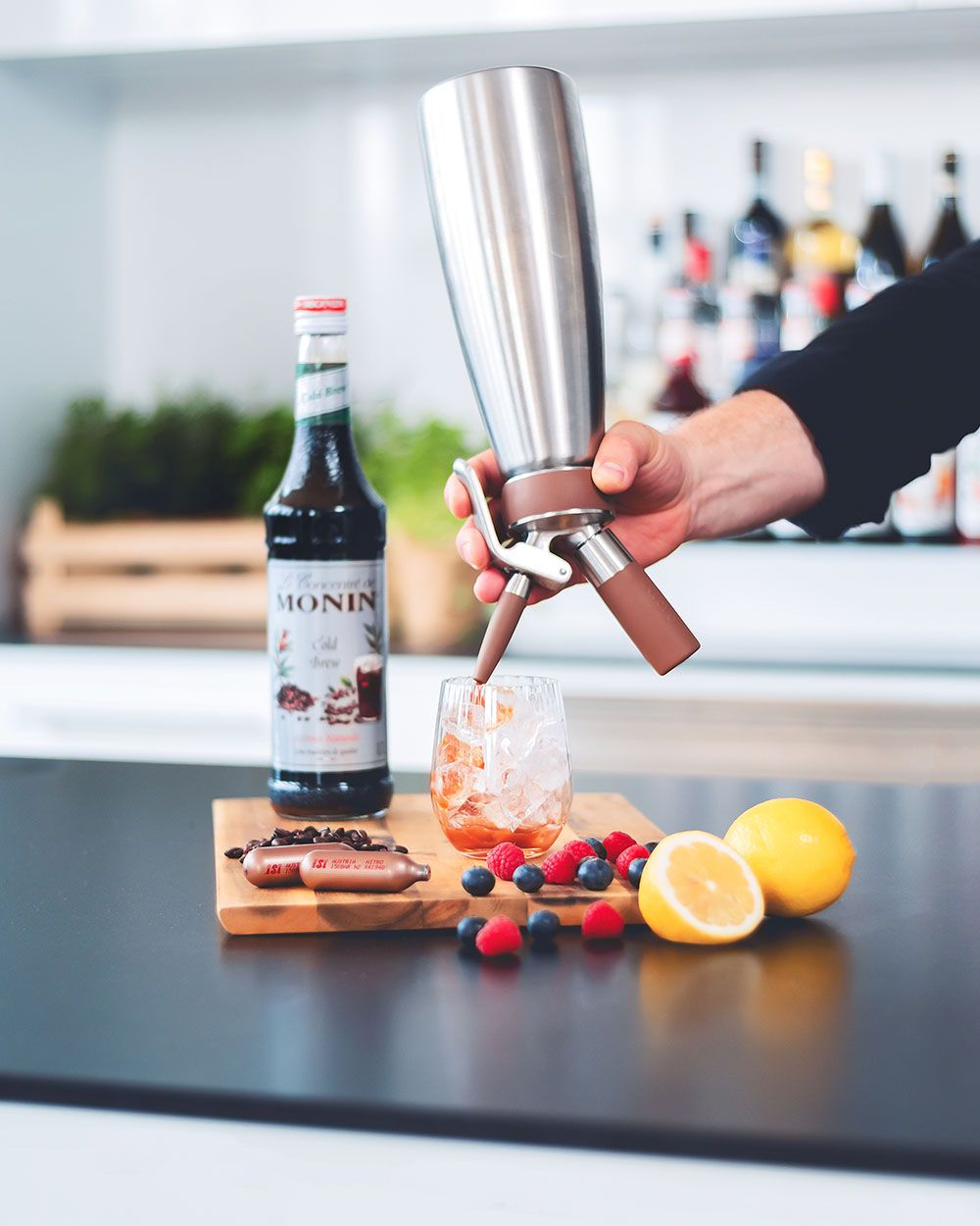 Jemand sprüht Bramble mit einem iSi Nitro in ein Glas, daneben Beeren, Zitronen, Monin Sirup und iSi Charge Kapseln. 