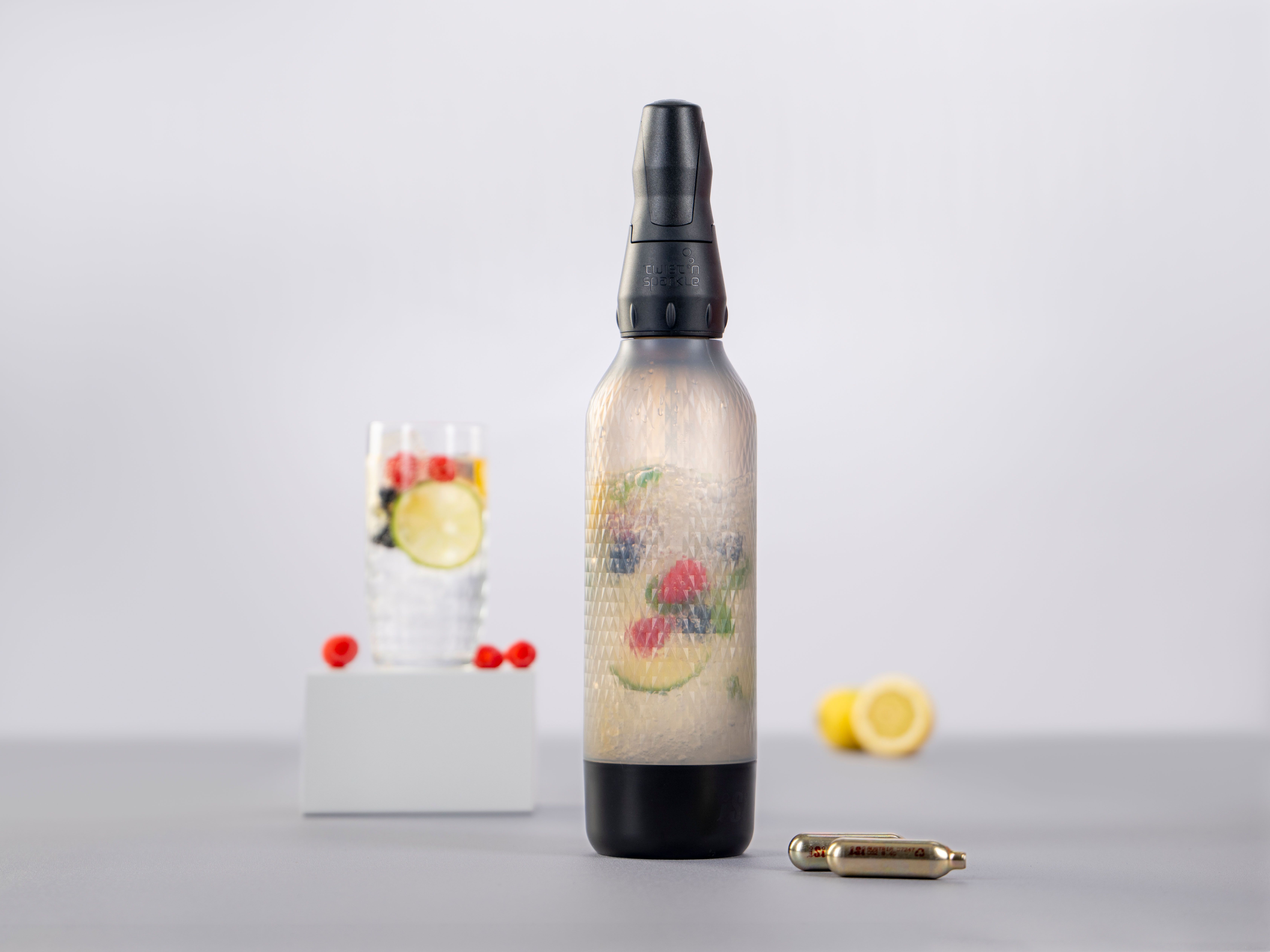 Ein Glas mit Sparkled Infused Wasser und Früchten, daneben weitere Früchte, ein iSi Twist'n Sparkle und iSi Charge Kapseln.