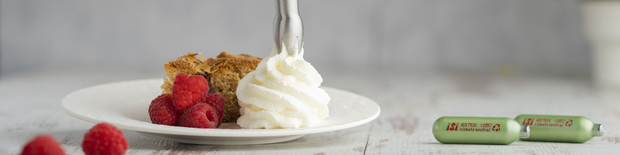 Auf einem Teller werden mit dem iSi Green Whip frische Schlagsahne neben Kuchenstück und Himbeeren aufgespritzt, daneben liegen zwei grüne iSi Kapseln.