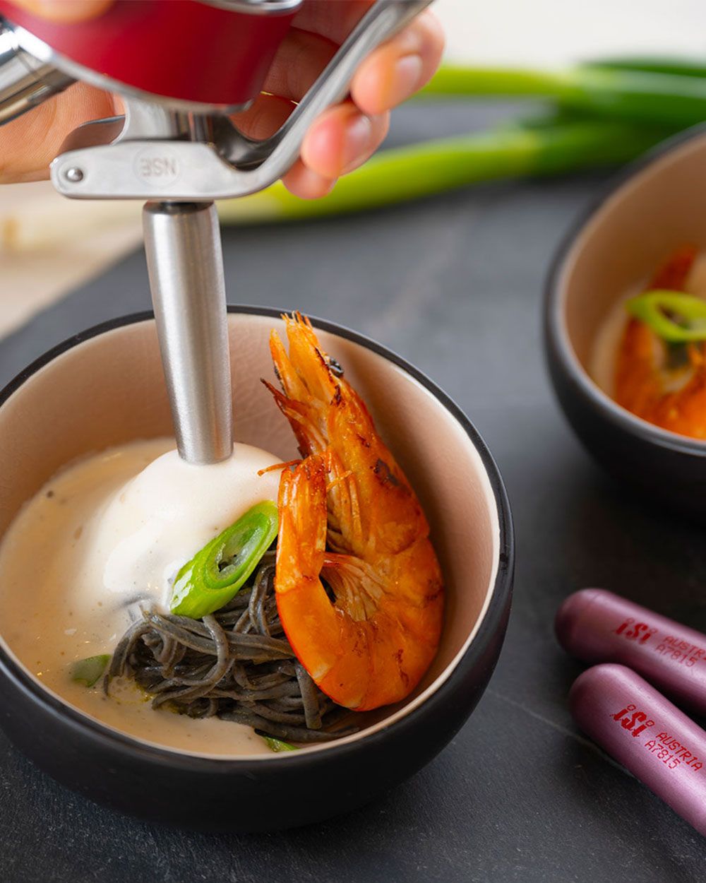 Jemand sprüht mit einem iSi Gourmet Whip Lachssauce in eine Schüssel mit Nudeln und einem Scampi, daneben iSi Charge Kapseln.
