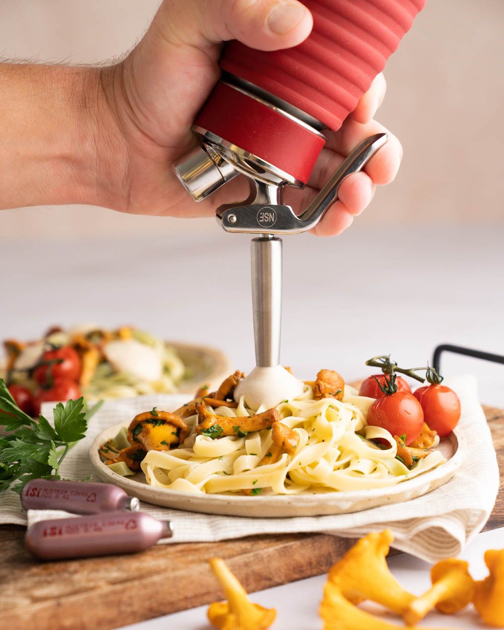 Jemand nutzt ein iSi Gourmet Whip, um Pilzrahmsauce auf einen Teller mit Pasta zu sprühen. Daneben liegen Tomaten, Pilze und zwei iSi Charge Kapseln. 