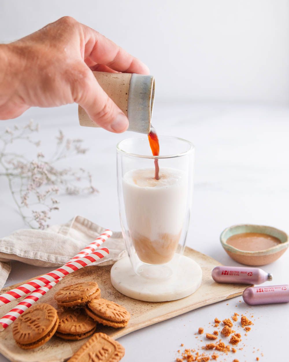 Jemand gießt Kaffee in ein Glas mit Milch, daneben Lotus-Kekse, Gewürze und zwei iSi Charge Kapseln. 