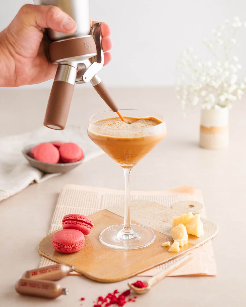 Jemand sprüht mit einem iSi Nitro Espresso Martini in ein Glas, daneben iSi Charge Kapseln und Macarons 