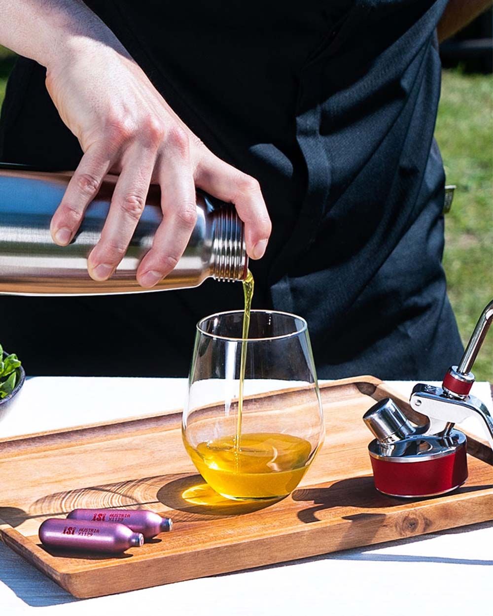 Jemand gießt aus einer iSi Gourmet Whip indisches Öl in ein Glas, daneben zwei iSi Charge Kapseln. 