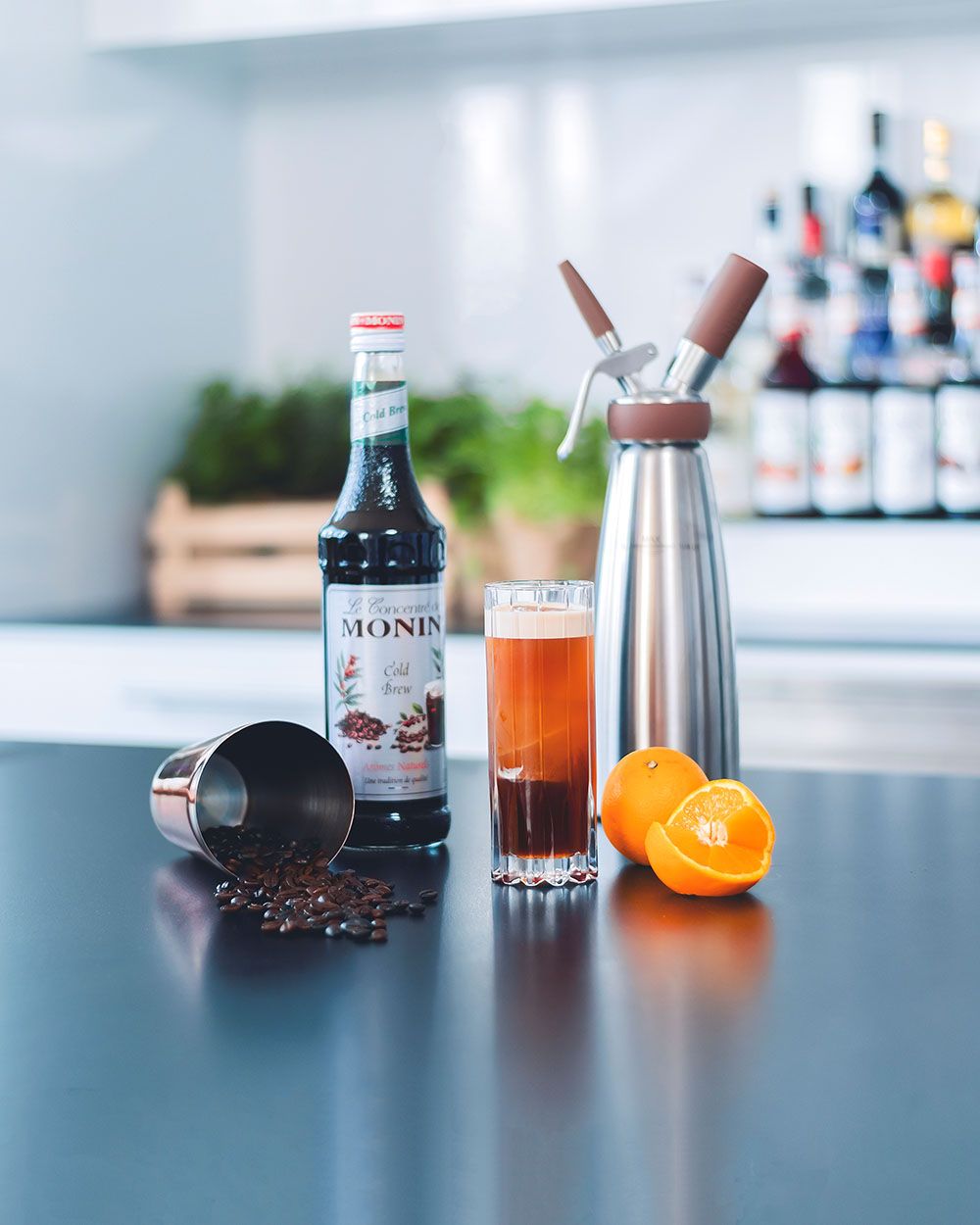 Ein Glas mit Fruity Tonic, daneben eine Flasche Sirup, eine Orange, Kaffeebohnen und ein iSi Nitro. 