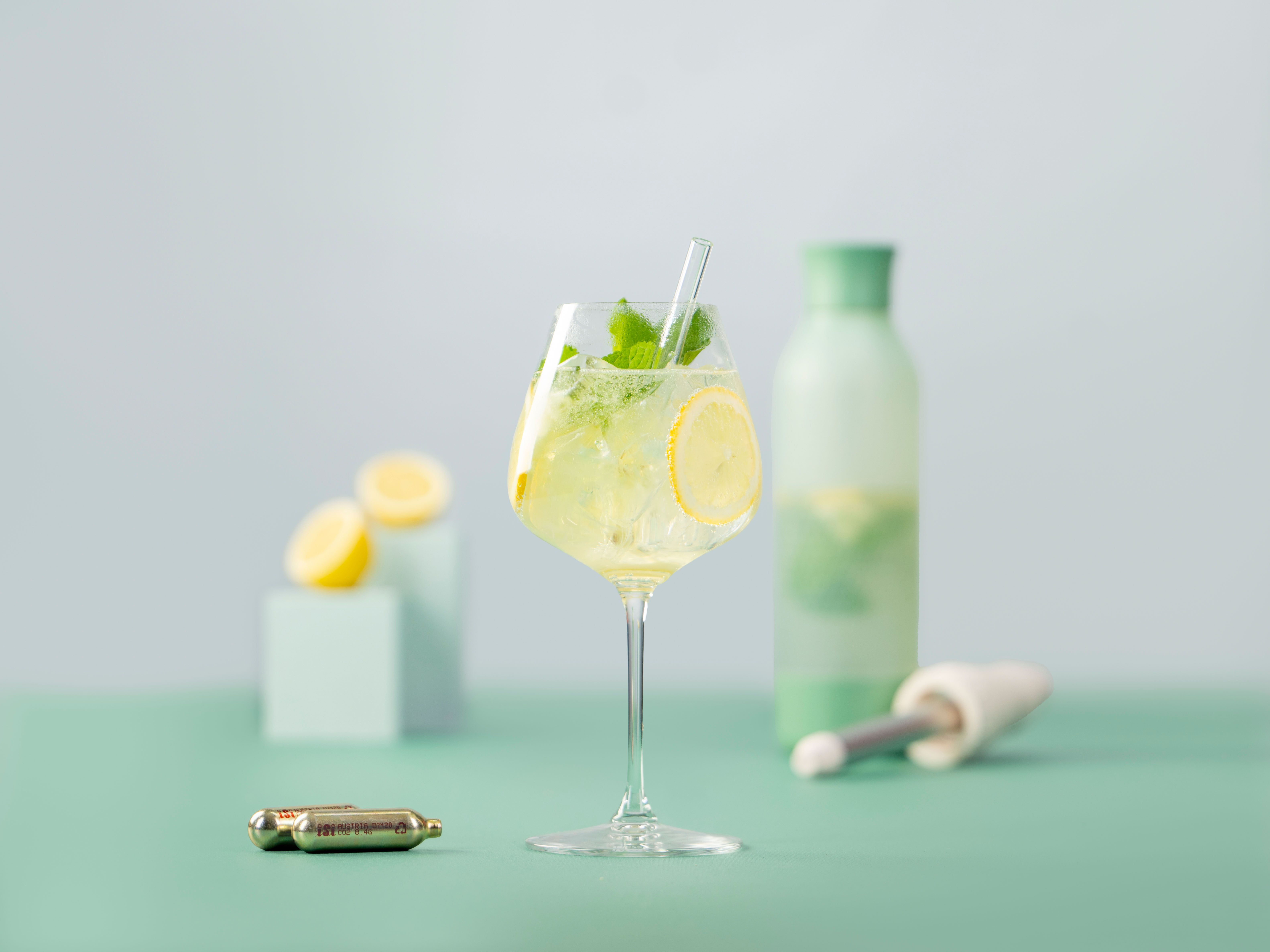 Ein Glas mit Hugo Bliss, daneben iSi Charge Kapseln, im Hintergrund Zitronen und ein iSi Twist'n Sparkle. 