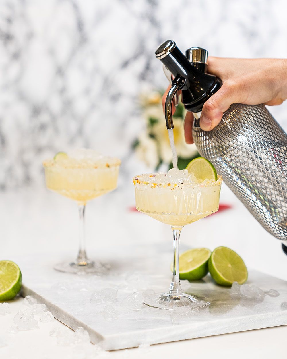 Jemand füllt aus einem iSi Sodamaker Soda in ein Glas mit Holunder-Minz-Limonade, daneben Limetten. 