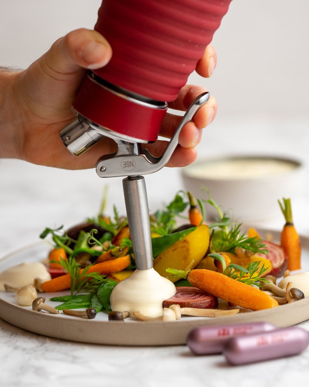 Jemand sprüht mit einem iSi Gourmet Whip Polentaschaum auf einen Teller mit Gemüse. 