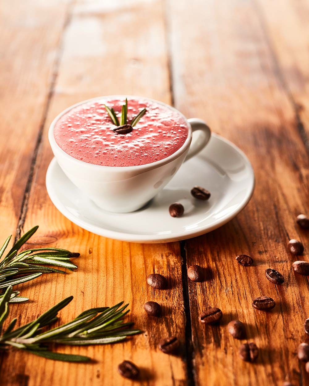 Eine weiße Tasse mit rosa Cremeschaum ist mit einem Rosmarinzweig und einer Kaffeebohne garniert und steht auf einer Untertasse, umgeben von Kaffeebohnen und Rosmarinzweigen.
