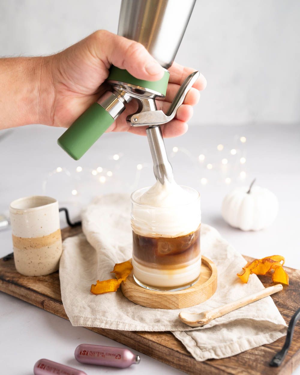 Jemand nutzt einen iSi Gourmet Whip, um Schaum auf ein Kaffeegetränk zu sprühen. Daneben Kürbis und iSi Charge Kapseln. 