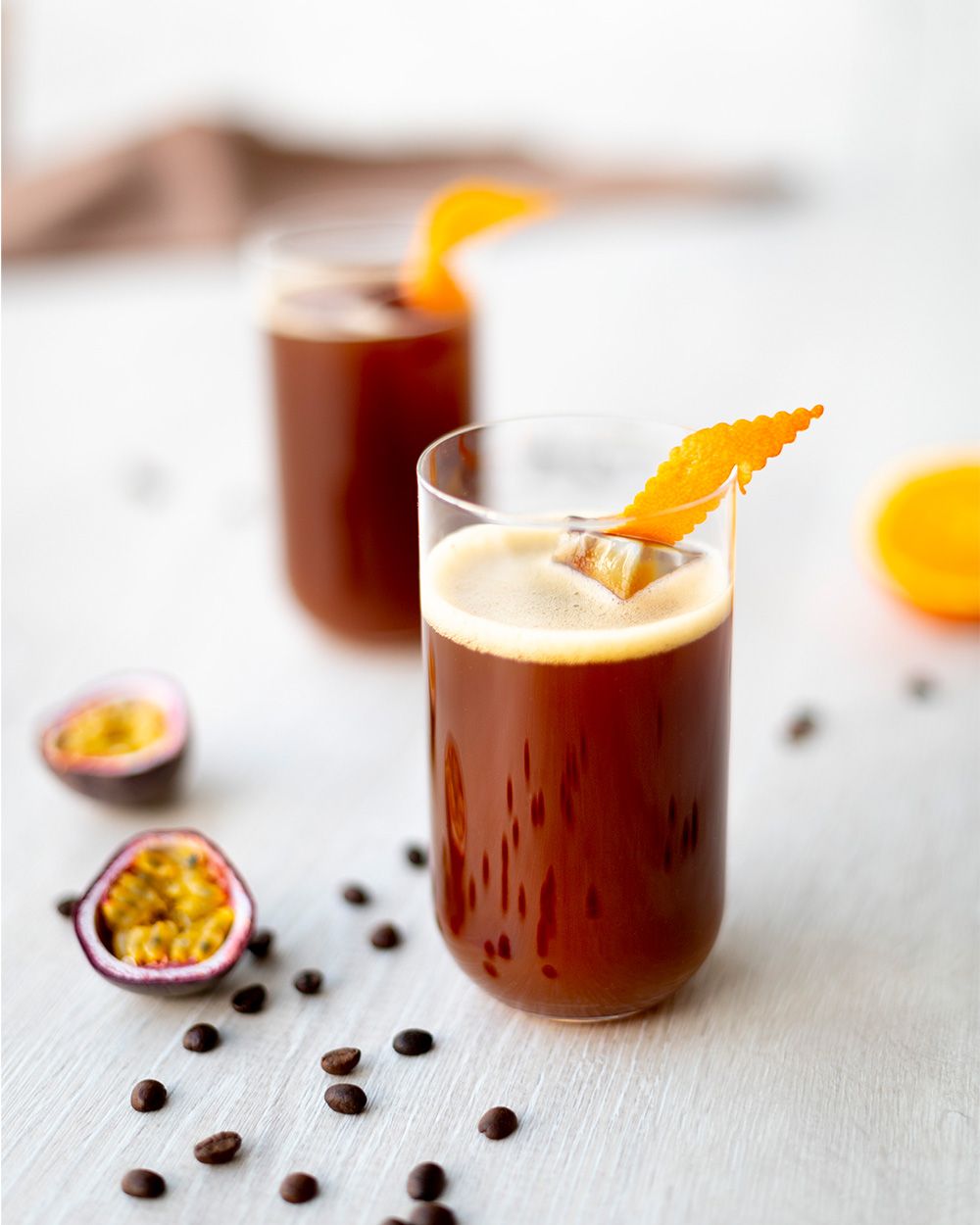 Zwei Gläser mit einem Kaffee-Orange-Getränk, daneben Passionsfrüchte und Kaffeebohnen. 