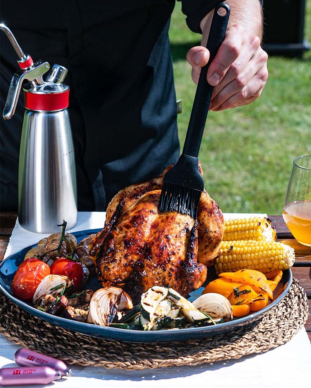 Jemand streicht indisches Öl auf ein Brathuhn, neben dem Gemüse auf dem Teller liegt. Im Hintergrund ein iSi Gourmet Whip und iSi Charge Kapseln. 