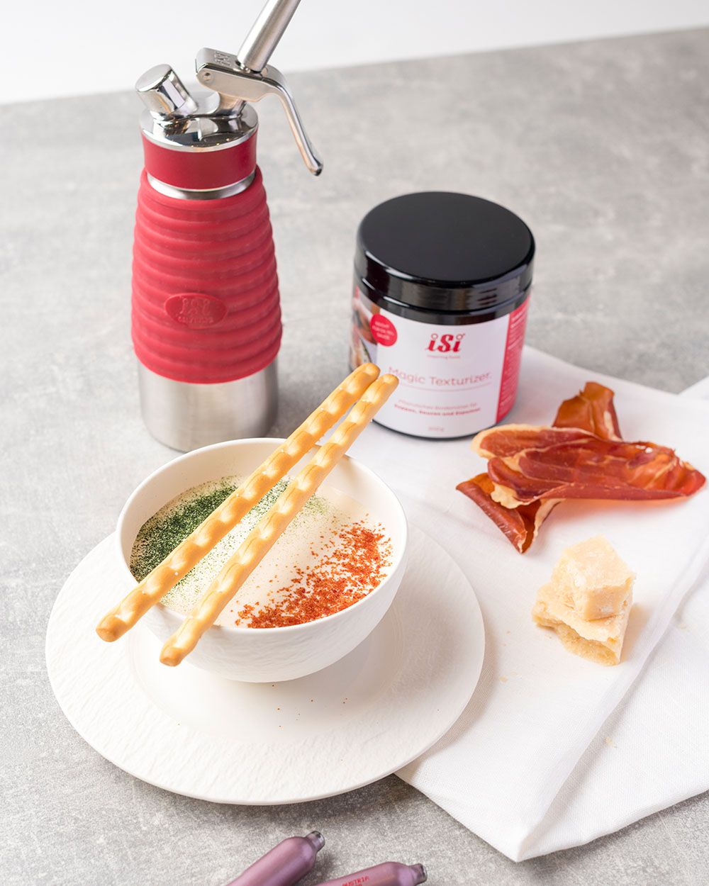 Eine Schüssel mit Parmesan Espuma, daneben Prosciutto, Parmesan, ein iSi Gourmet Whip und iSi Magic Texturizer. 