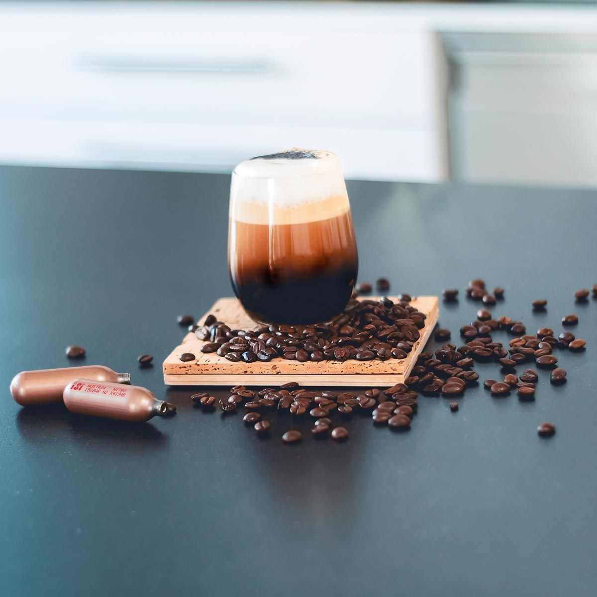 Ein Kaffeegetränk mit Schaum auf einem Brett mit Kaffeebohnen, daneben liegen zwei iSi Charge Kapseln. 
