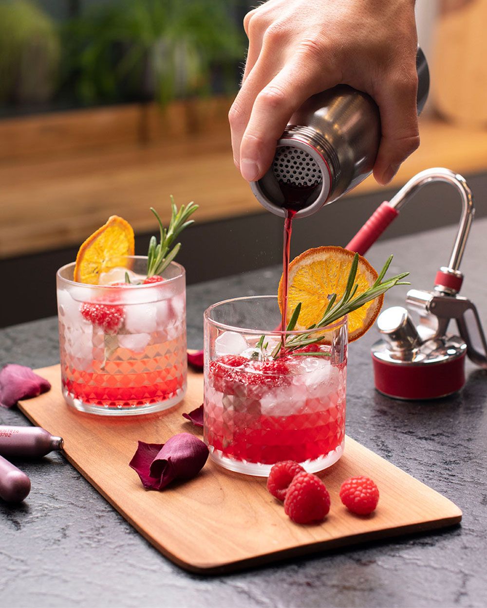 Jemand gießt Himbeer-Sirup aus einem iSi Gourmet Whip in ein Glas, daneben ein weiteres Glas mit einem roten Getränk und iSi Charge Kapseln. 