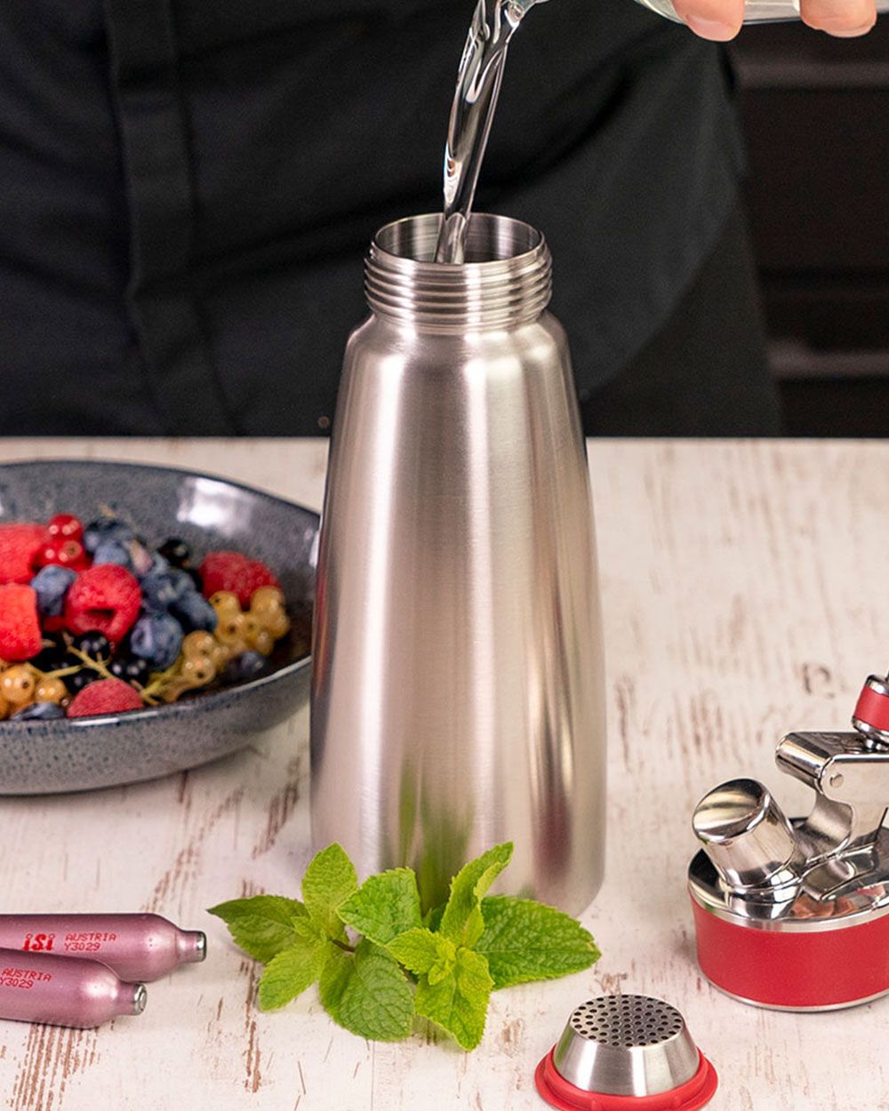 Jemand gießt Wasser in einen Gourmet Whip, daneben liegen zwei iSi Charge Kapsel, Minze, ein Teller mit Beeren und Müsli und ein Krug mit Wasser. 