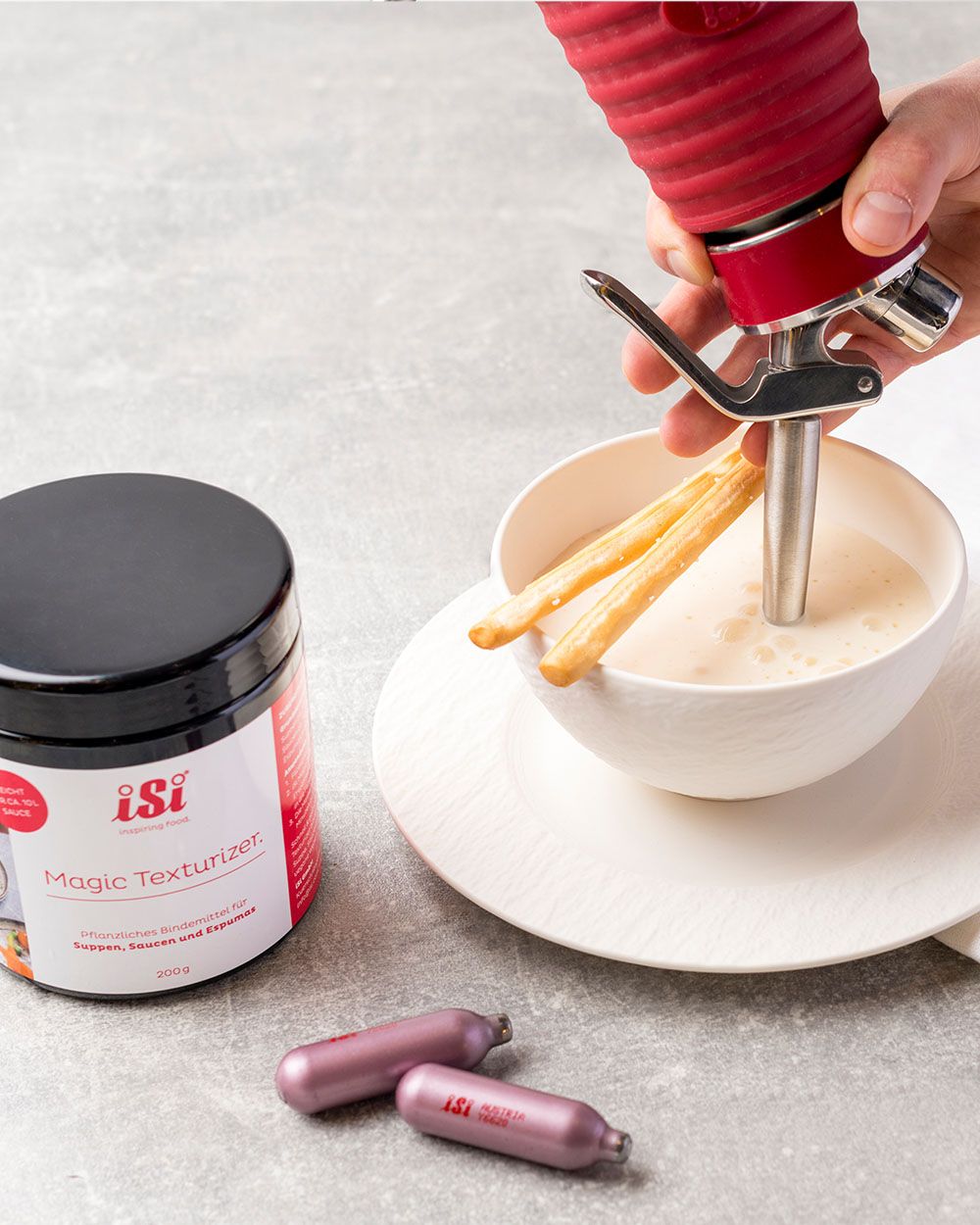 Eine Hand spritzt mit einem iSi Whip cremige Suppe in eine Schale mit Brotstangen, daneben stehen iSi Magic Texturizer und zwei iSi Kapseln.