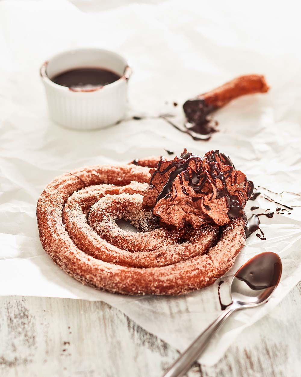 Eingerollte Churros mit Schokoladeschaum, daneben ein Löffel und im Hintergrund eine Schüssel mit Schokosauce. 