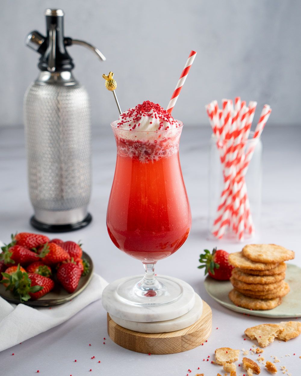 Ein roter Erdbeer-Drink im Glas mit Sahnehaube, Strohhalm und Streuseln steht auf einem Tisch, umgeben von frischen Erdbeeren, Keksen, einem iSi Sodamaker und roten Papierstrohhalmen.