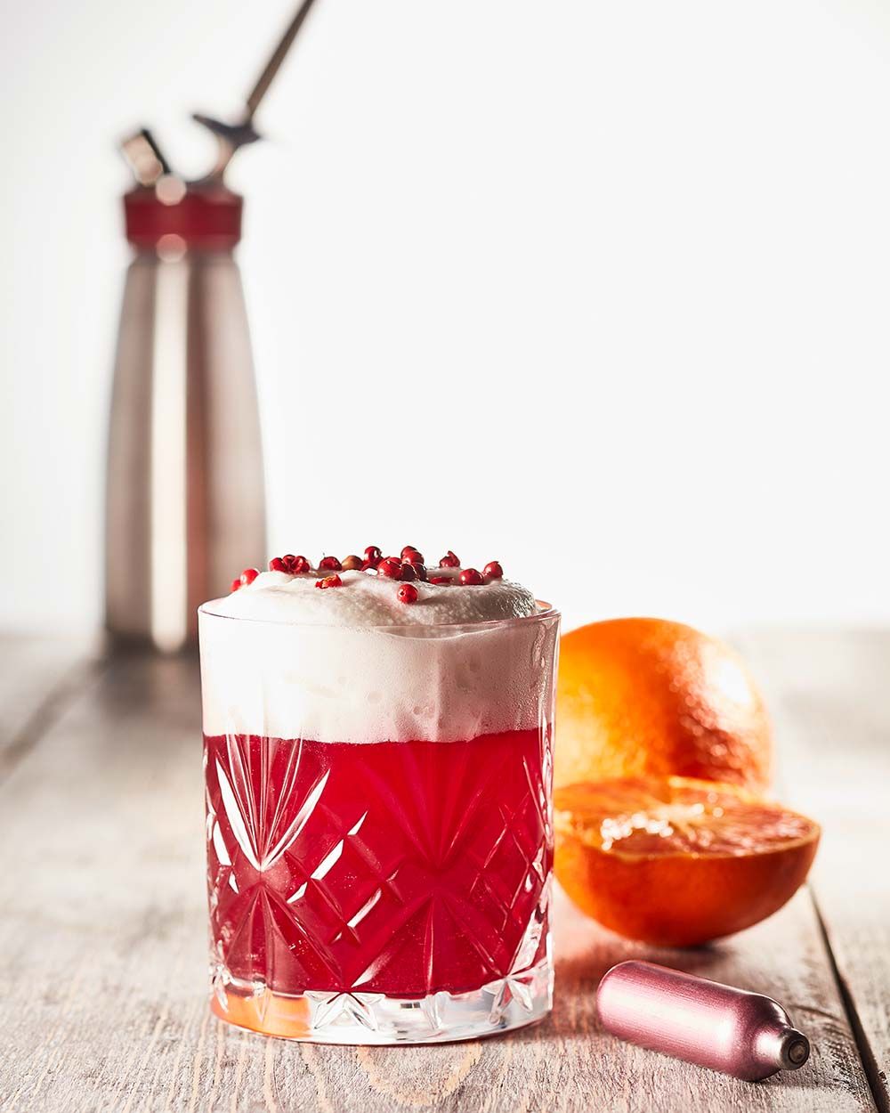 Ein rotes Getränk mit Schaum, daneben eine iSi Charge Kapsel und eine Orange, im Hintergrund ein Gourmet Whip.