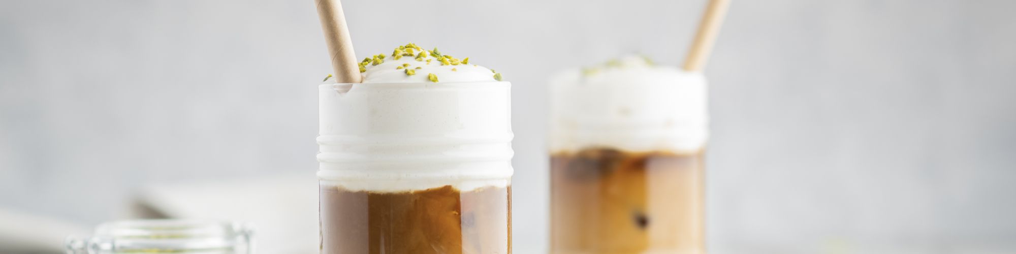 Zwei Gläser mit Eiskaffee und dicker Schaumhaube sind mit gehackten Pistazien bestreut und jeweils mit einem Papierstrohhalm serviert.