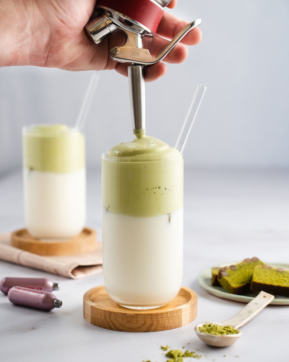 Jemand sprüht Matcha-Topping auf ein Milchgetränk, daneben Matchapulver und iSi Charge Kapseln. 