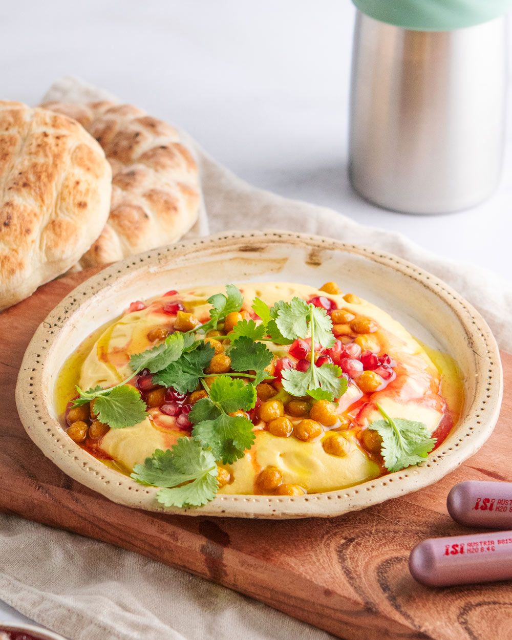 Ein Teller mit Kürbishummus mit Kichererbsen und Petersilie, daneben Pitabrot und zwei iSi Charge Kapseln. Im Hintergrund ein iSi Creative Whip. 