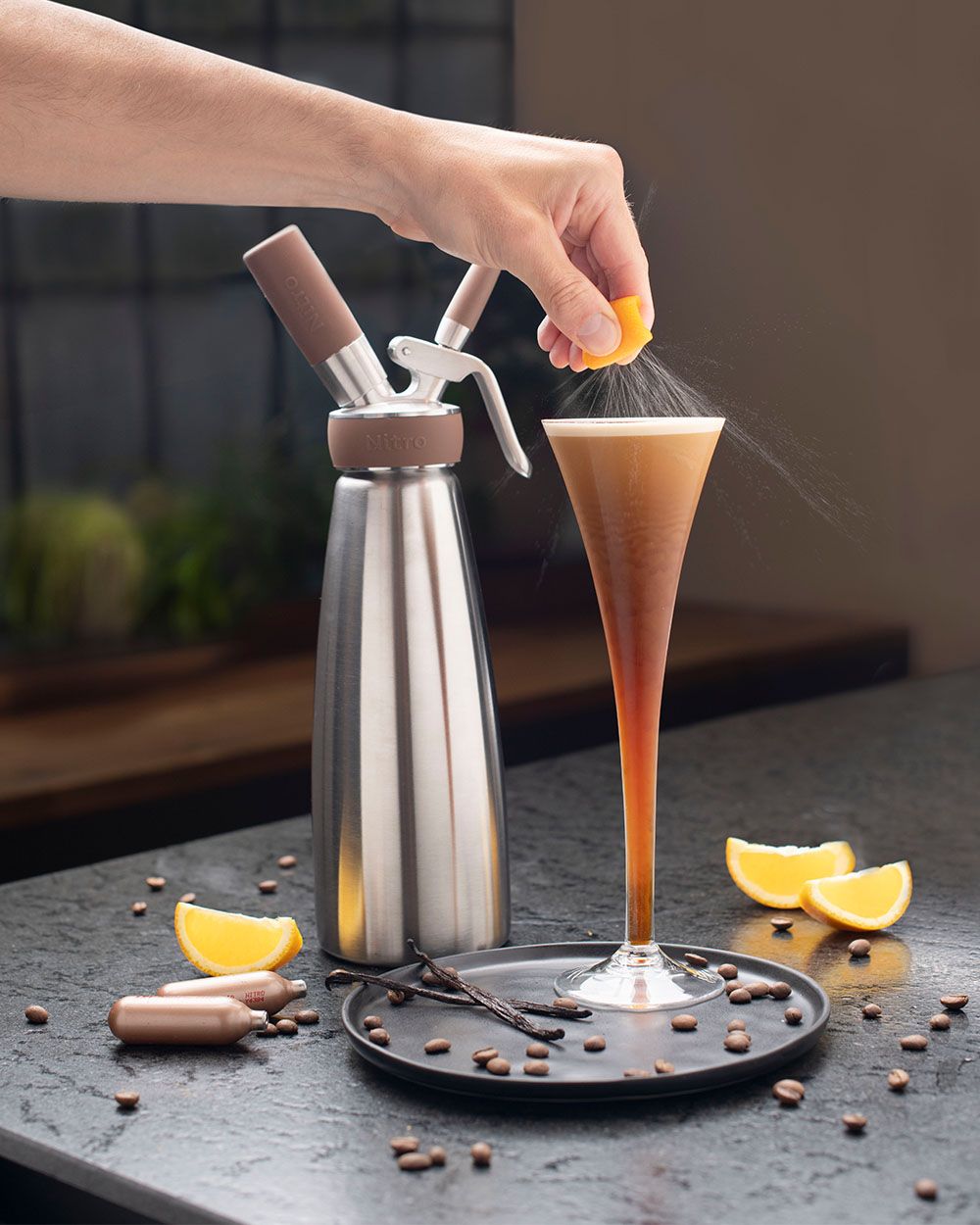 Jemand presst eine Orange in ein Glas mit Nitro Espresso Martini, daneben Kaffeebohnen, Orangen und ein iSi Nitro. 