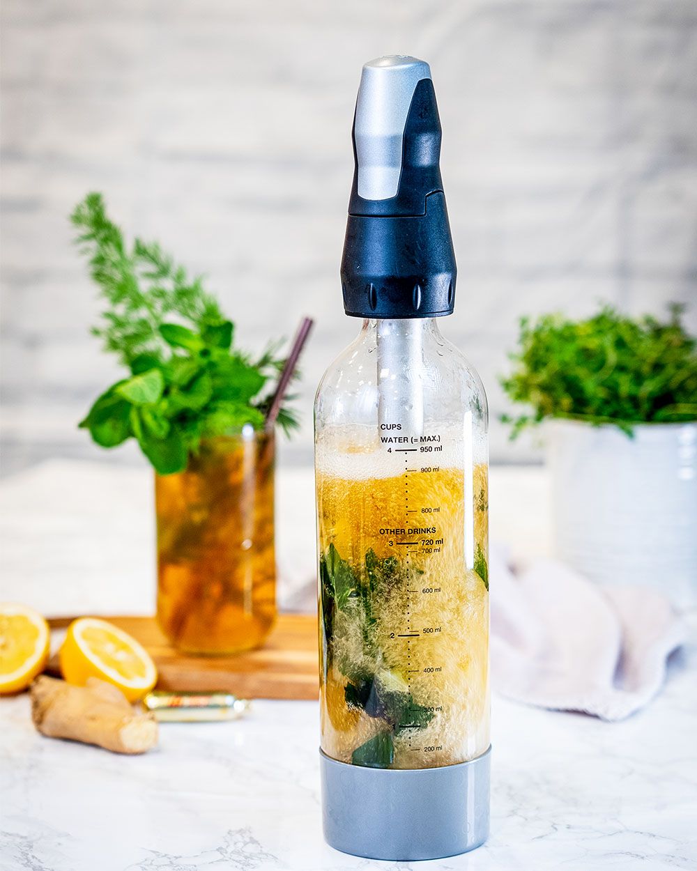 Ein Eistee Getränk in einem iSi Sodamaker, im Hintergrund ein Glas mit dem fertigen Getränk, Zitronen und Ingweg. 