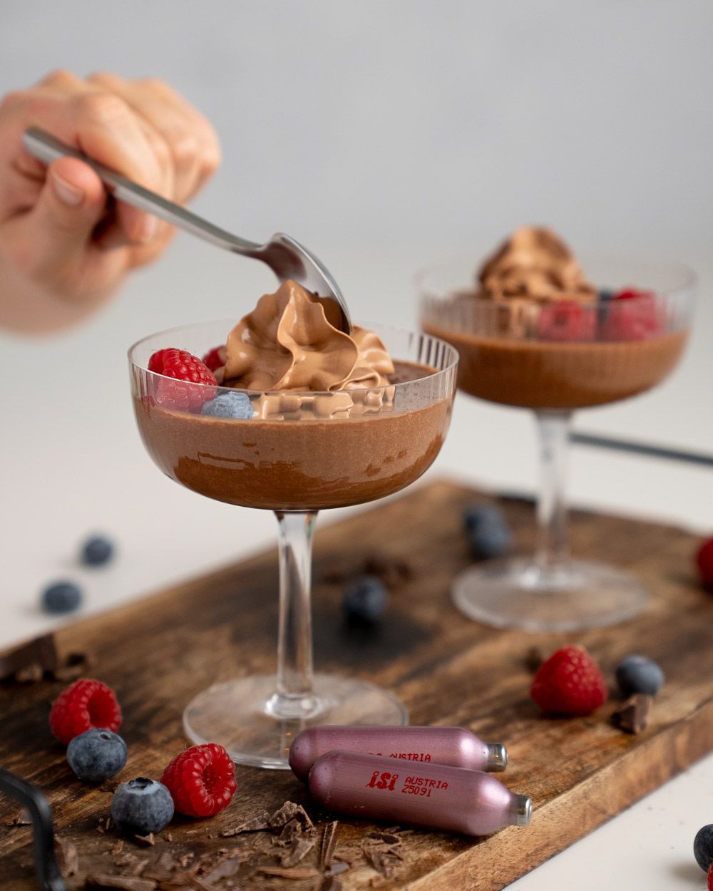 Jemand fährt mit einem Löffel in ein veganes Schokomousse, daneben frische Beeren, iSi Charge Kapseln und ein weiteres Glas mit Mousse. 