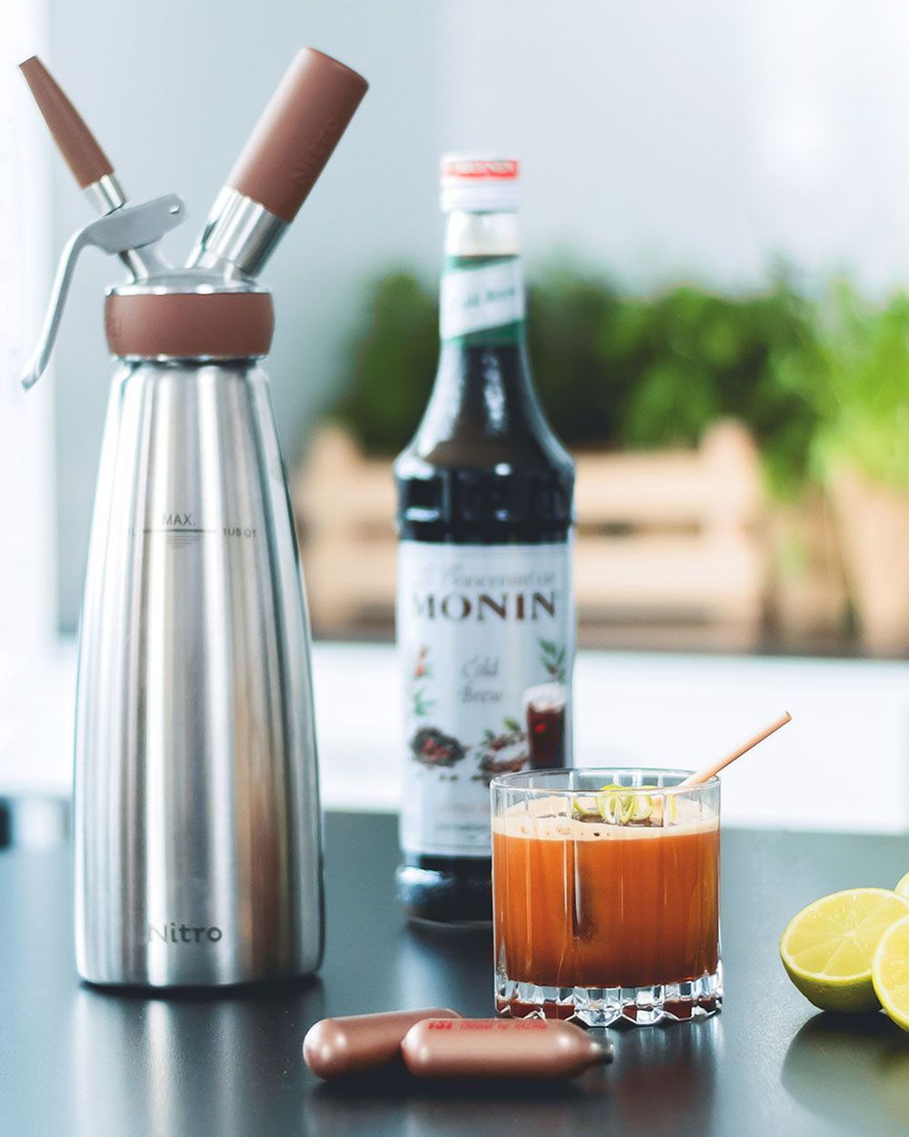 Ein Kaffee-Gin-Getränk, daneben iSi Charge Kapseln, eine Limette, eine Flasche Monin Sirup und eine iSi Nitro. 