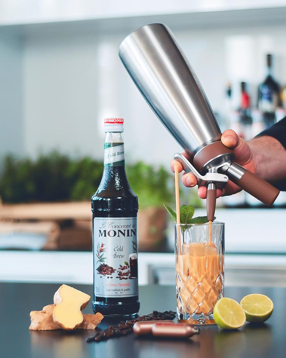 Jemand sprüht mit einem iSi Nitro Ginger-Lemon Coldbrew in ein Glas, daneben iSi Charge Kapseln, Limetten, Ingwer und eine Flasche Monin Sirup.