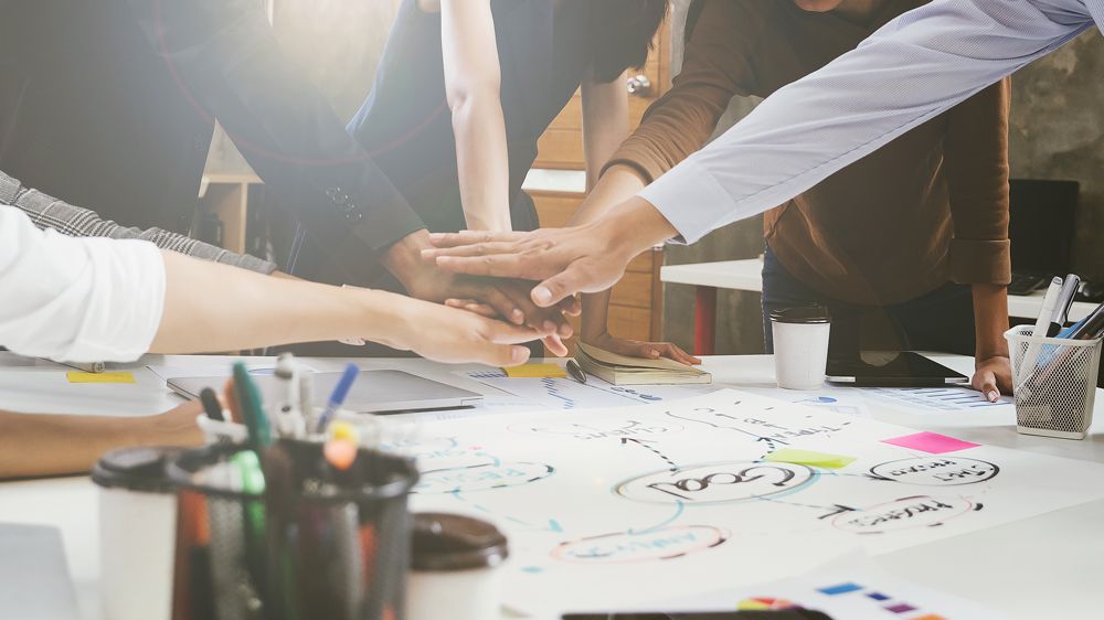 Mehrere Personen legen ihre Hände gemeinsam auf einen Tisch, auf dem Skizzen, Notizen und Marker liegen, als Zeichen von Teamarbeit.