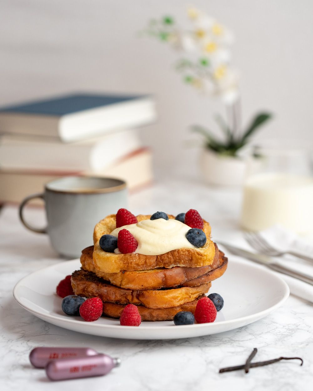Ein Teller mit French Toast ist mit Beeren und cremigem Topping garniert, daneben liegen iSi Kapseln und eine Vanilleschote, im Hintergrund stehen Bücher, eine Tasse Kaffee und ein Glas Milch.